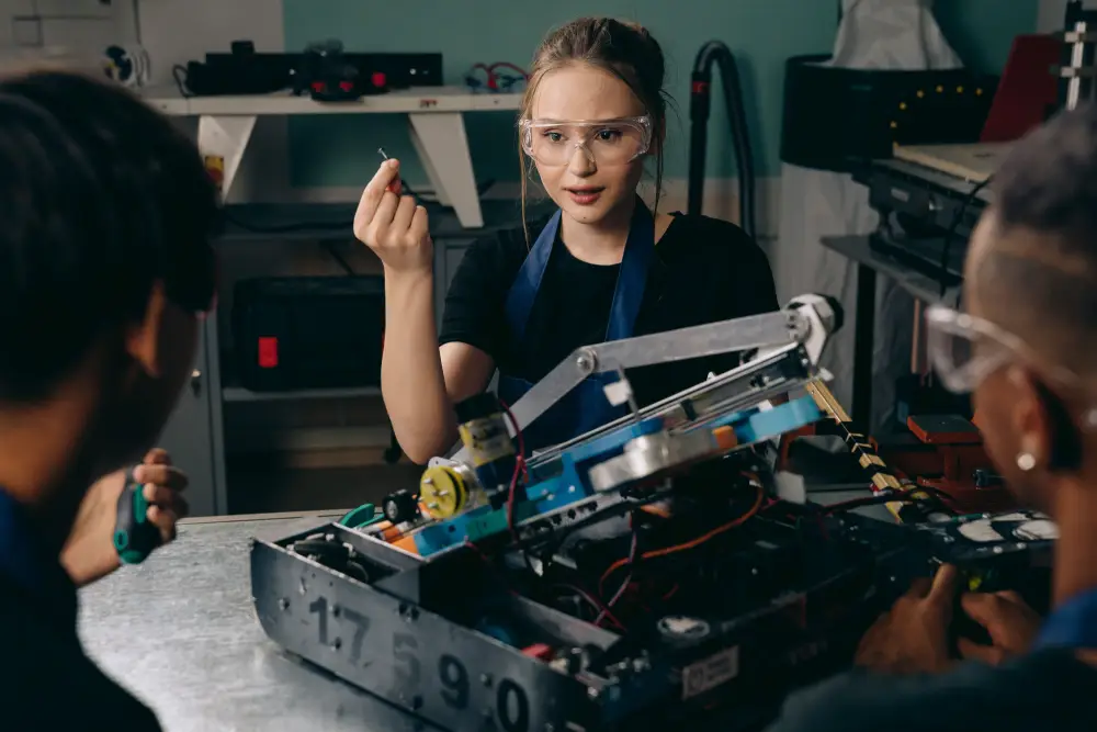 engineering-students-robotics-workshop A team of engineering students wearing safety glasses working on a custom robotics chassis in a workshop, while a female student holds a small screw and explains the project.