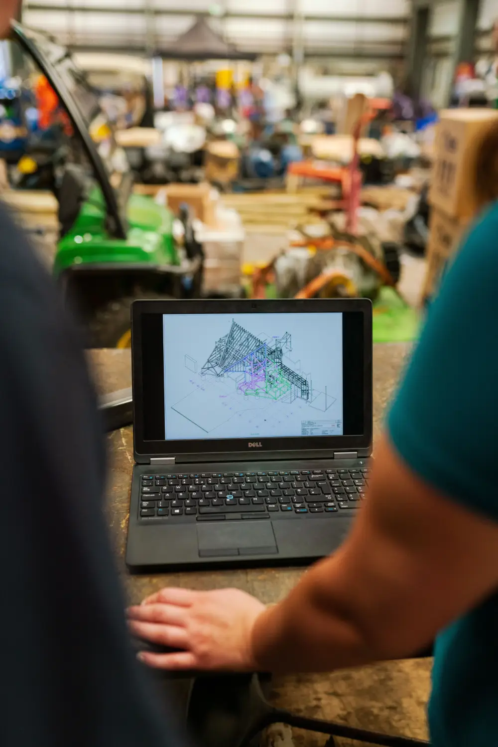 A close-up shot of a person using a Dell laptop in an industrial workshop to view a 3D structural CAD model, with machinery blurred in the background.